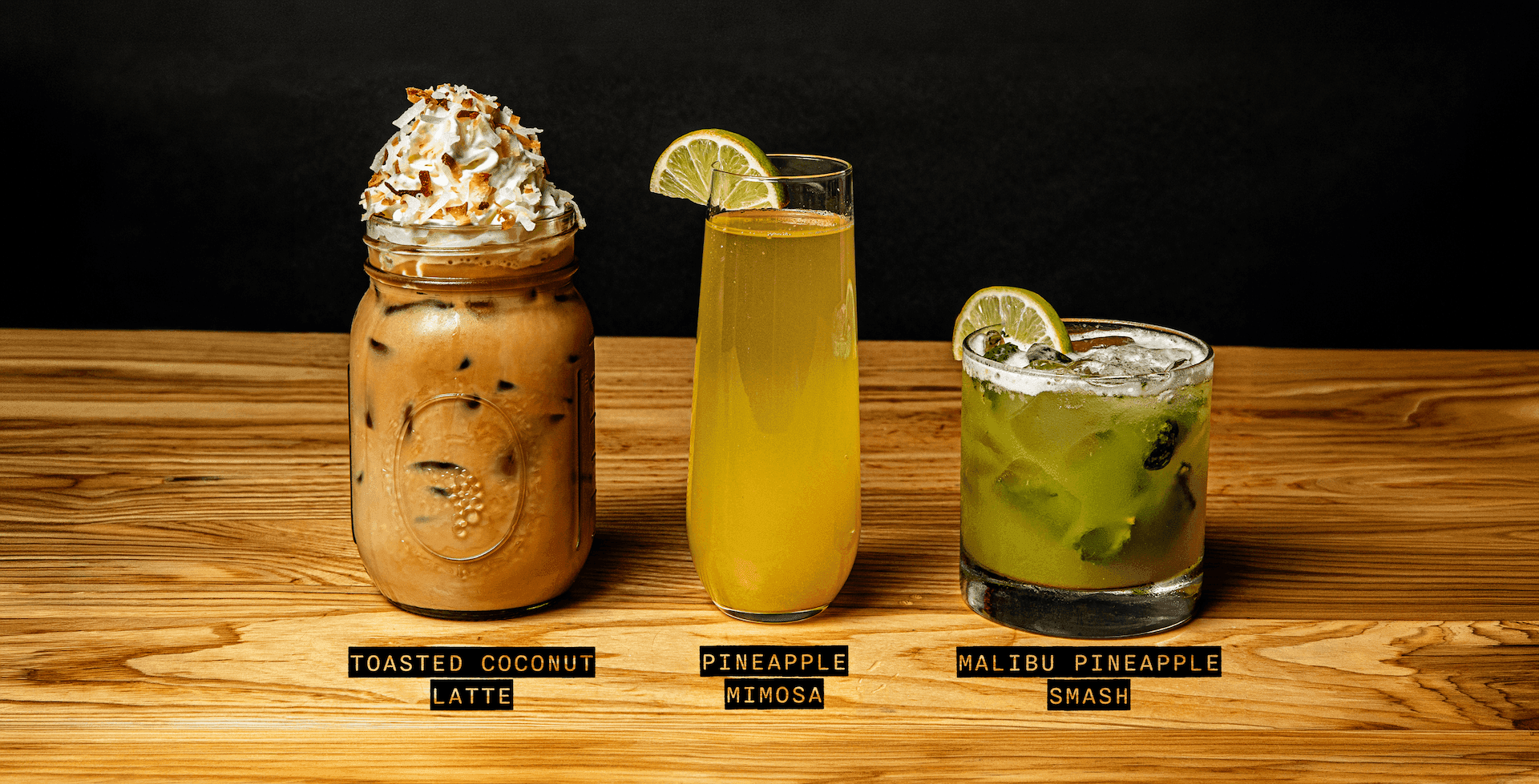 Three drinks on a wooden table: a topped iced latte in a jar, a yellow mimosa in a flute with a lime wedge, and a green cocktail in a rocks glass with a lime wedge. Labels identify each drink.