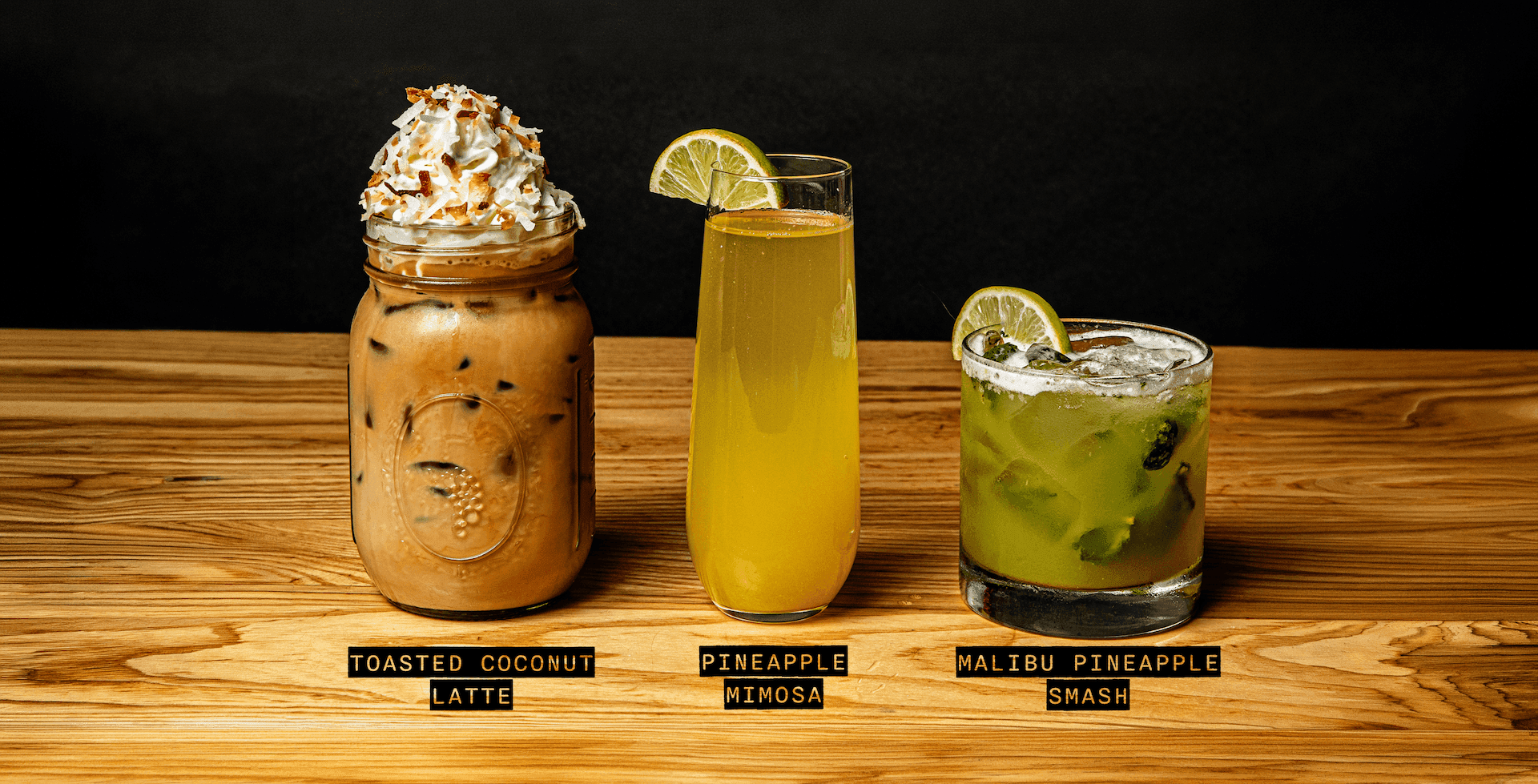 Three drinks on a wooden surface: a toasted coconut latte with whipped cream, a pineapple mimosa with a lime wedge, and a malibu pineapple smash with ice and a lime wedge.