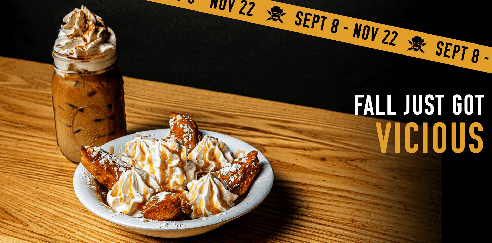 A mason jar with whipped cream next to a plate of fried pastries drizzled with caramel sauce and powdered sugar on a wooden table. Text reads, FALL JUST GOT VICIOUS..