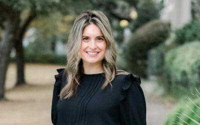 A woman with blonde hair, wearing a black long-sleeve top, stands outdoors smiling at the camera with trees and greenery in the background, ready to enjoy a brunch full of flavor.