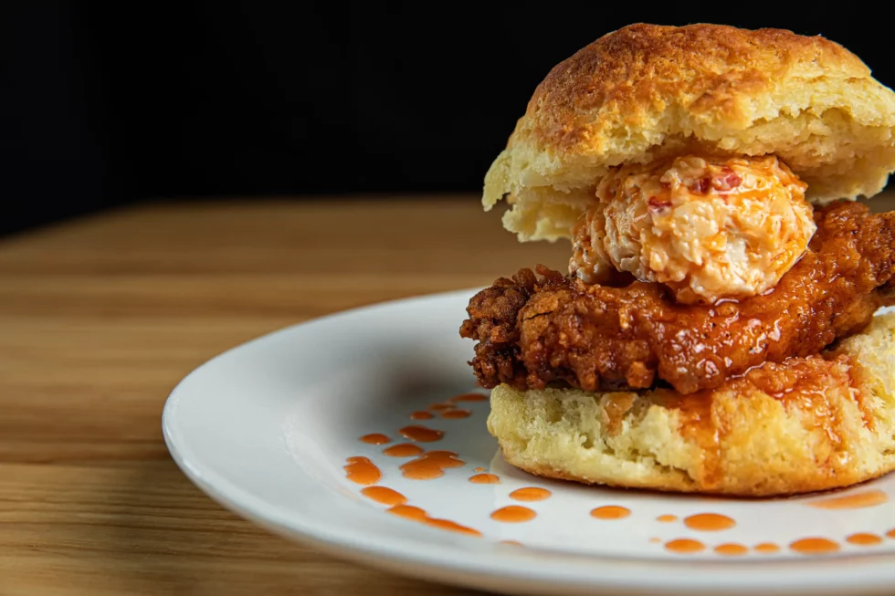 A fried chicken biscuit sandwich full of flavor is topped with a dollop of pimento cheese and drizzled with hot sauce, perfect for breakfast or brunch.