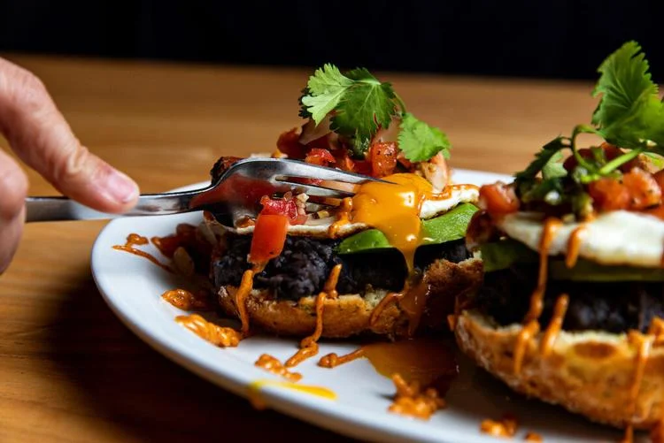 A fork cuts into a runny egg atop black beans, avocado, salsa, and sauce on toasted bread, garnished with cilantro. This delicious breakfast is inspired by Vicious Biscuit’s take on Southern cooking.
