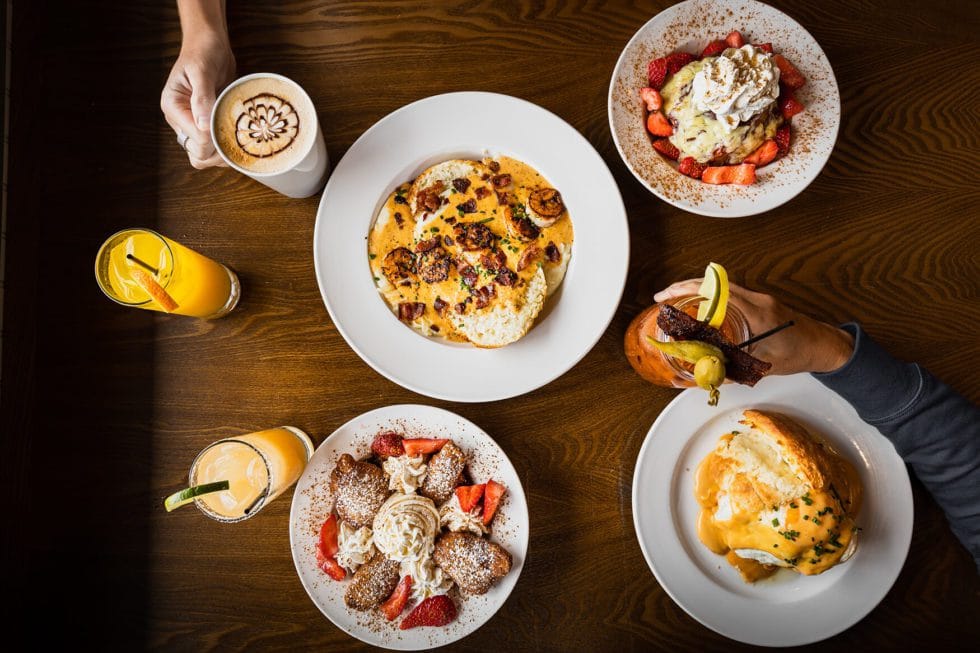 A table full of flavor features breakfast dishes like eggs, pancakes with strawberries, beignets, biscuits from Vicious Biscuit, two orange drinks, and coffee with latte art. Two hands reach for the drinks.