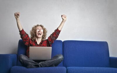 A person with curly hair sits on a blue sofa, raising both arms in excitement while looking at a laptop, ready to enjoy breakfast full of flavor.