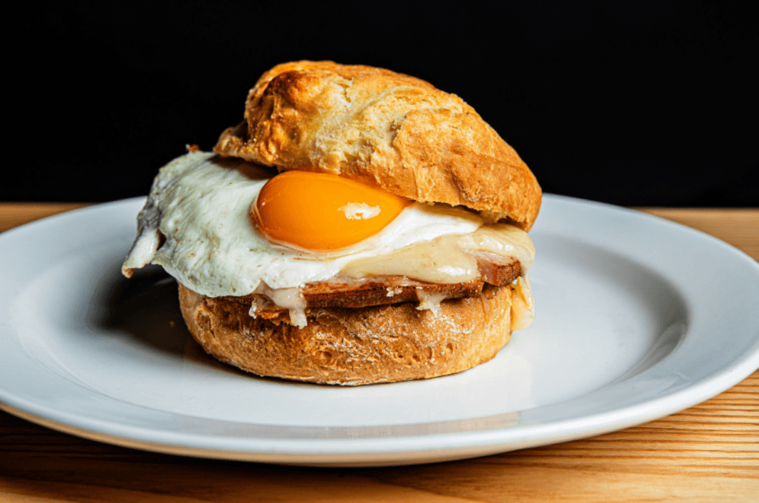 A delicious breakfast sandwich with a fried egg, melted cheese, and meat on a round bun is served on a white plate.