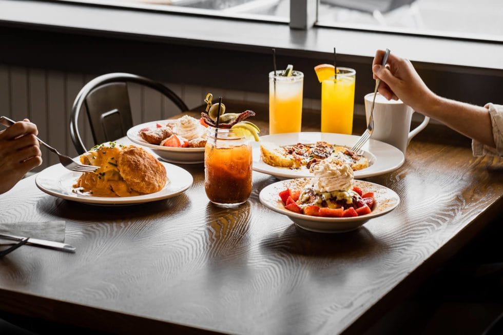 Two people enjoy a Vicious Biscuit breakfast at a wooden table, savoring omelets, waffles with strawberries and whipped cream, drinks, and a Bloody Mary in a bright setting—full of flavor.