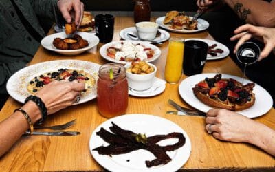Four people enjoy a breakfast full of flavor around a wooden table, savoring waffles, pancakes, fruit, bacon, sausage, juice, and coffee.