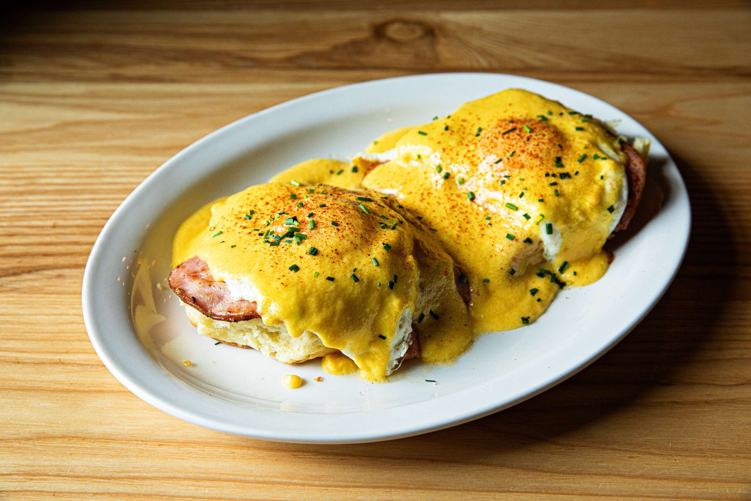Two servings of delicious eggs Benedict topped with hollandaise sauce and sprinkled with herbs are served on a white oval plate, perfect for breakfast or brunch.