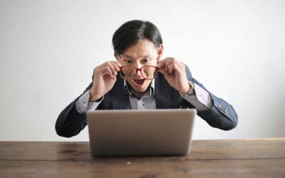 A man in a suit looks surprised while lifting his glasses and staring at a laptop on a wooden table, as if discovering something delicious and full of flavor.