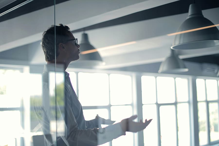 A person stands and gestures with hands in a modern office space, viewed through a glass wall with hanging lights overhead, creating an atmosphere as inviting as Southern Cooking.