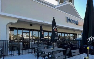 Outdoor seating area with black tables, chairs, and umbrellas in front of Victor’s, a restaurant serving delicious Southern cooking in a strip mall setting.