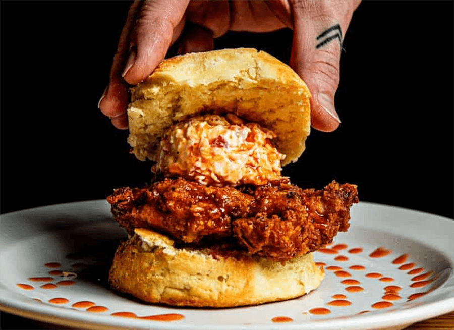 A hand holds a Vicious Biscuit sandwich filled with fried chicken and coleslaw, on a white plate decorated with dots of hot sauce—a true taste of Southern Cooking.