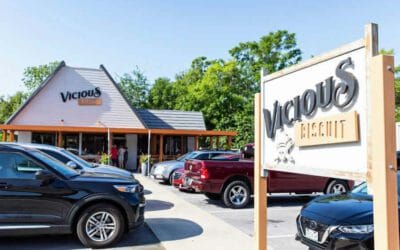 Several cars are parked outside Vicious Biscuit, with a large sign displaying the restaurant’s name in the parking lot. The spot is known for its delicious Southern cooking, and trees are visible in the background.
