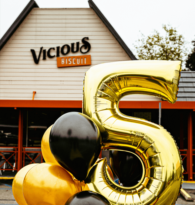 A gold number 5 balloon and black balloons are displayed outside a building with a sign that reads Vicious Biscuit, inviting you to enjoy delicious Southern cooking and brunch.