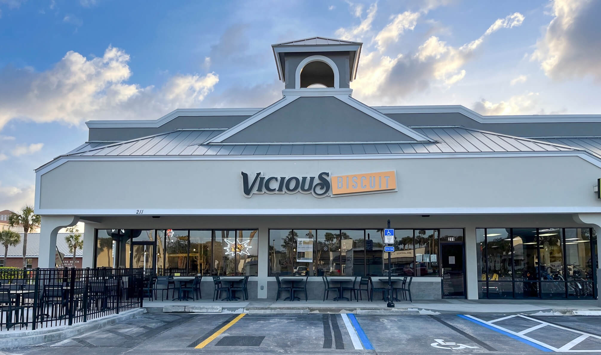 Exterior view of Vicious Biscuit restaurant with outdoor seating and glass windows, offering a delicious Southern cooking brunch experience. Visible parking spaces, including accessible spots, sit under a partly cloudy sky.