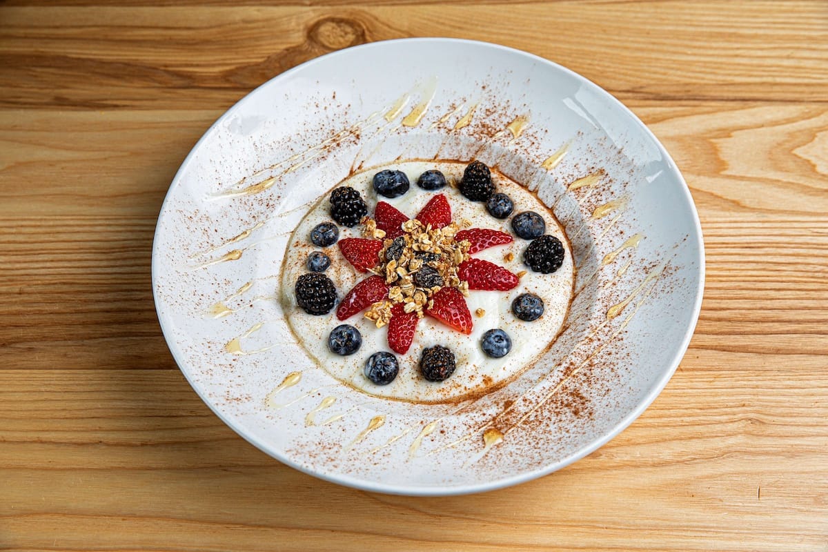 A white bowl with delicious yogurt topped with granola, blueberries, blackberries, sliced strawberries, honey drizzle, and a dusting of cinnamon sits on a wooden table—full of flavor like something from Vicious Biscuit.