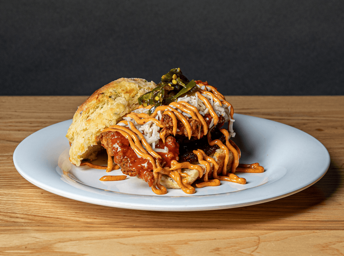 A Vicious Biscuit fried chicken biscuit sandwich topped with pickles and drizzled with orange and white sauces, served on a white plate on a wooden table—this breakfast is full of flavor.