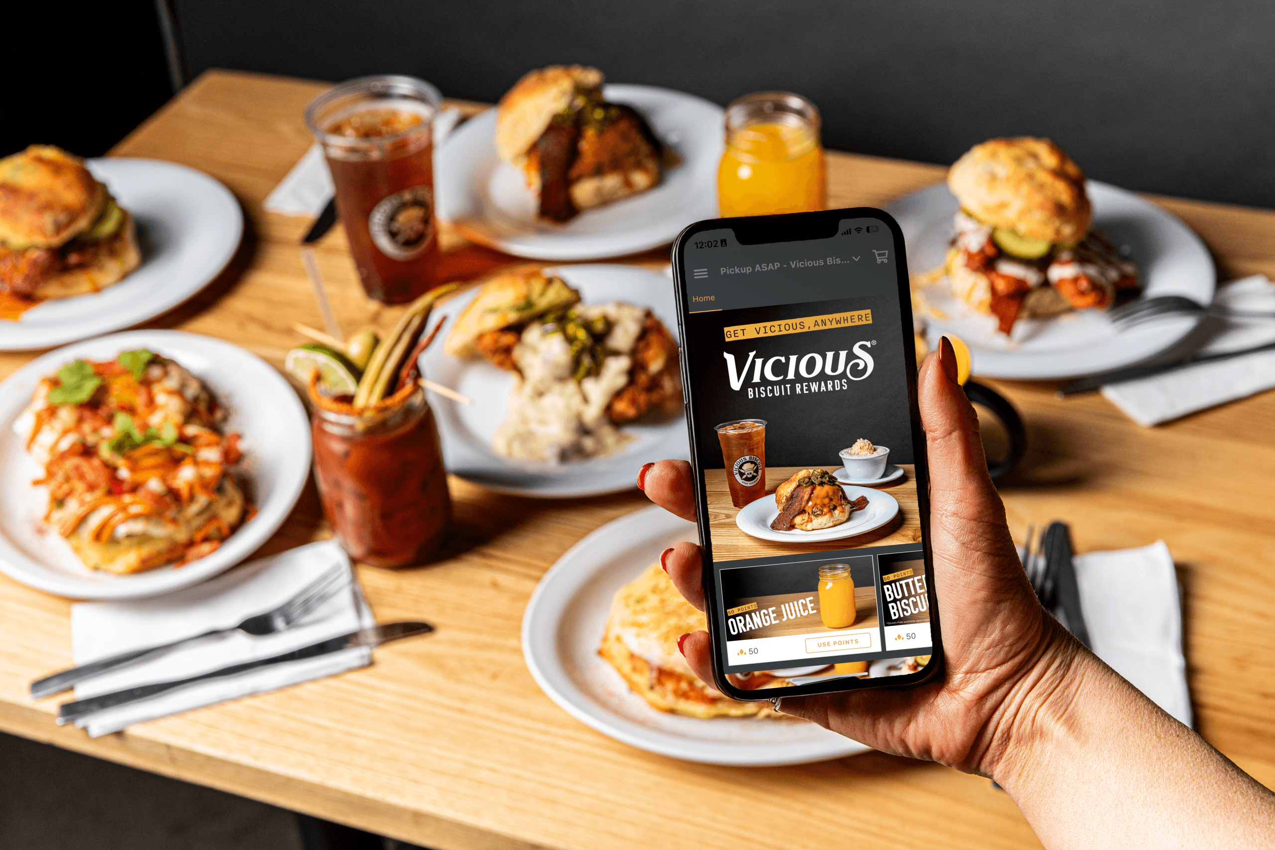 A hand holds a smartphone displaying the Vicious Biscuit menu over a table set with brunch plates, breakfast dishes, drinks, and cutlery—all full of flavor.