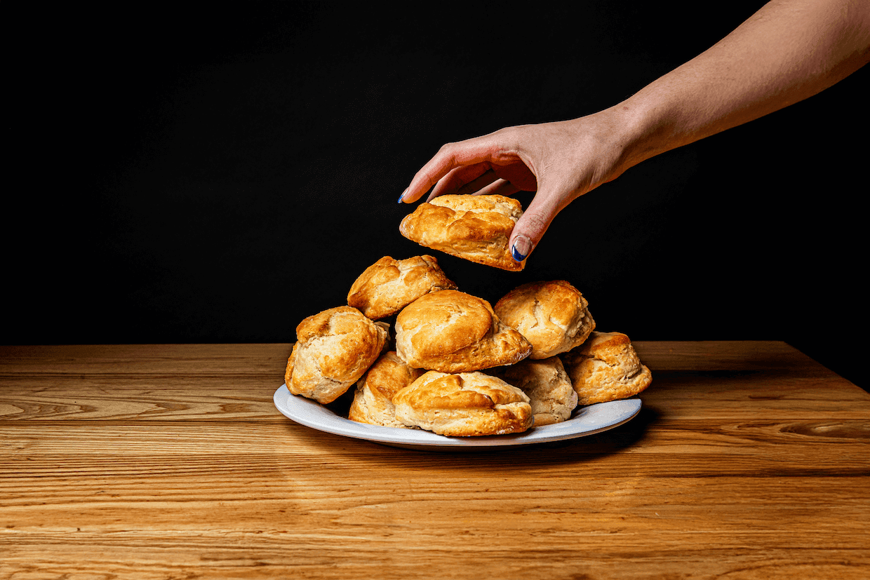A hand reaches to pick up a biscuit from a plate stacked with golden brown biscuits on a wooden table, capturing the essence of Southern cooking and breakfast, set against a black background.