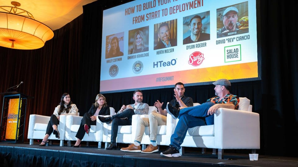 Five panelists sit on stage in white chairs, discussing a topic about building a brand, with a presentation slide and their photos displayed behind them at a conference.