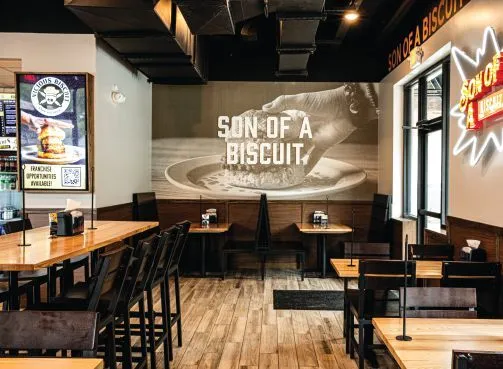 Interior of a restaurant with wooden tables and chairs, a mural reading Son of a Biscuit, and a neon sign on the wall.
