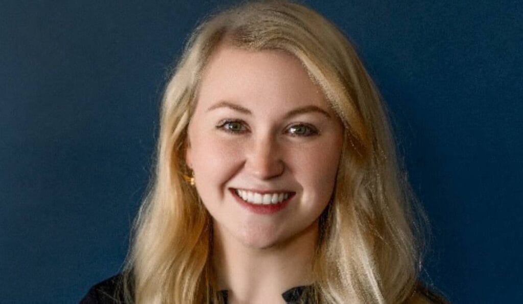 A woman with long blonde hair and light skin is smiling at the camera in front of a plain dark blue background. She is wearing a black top.