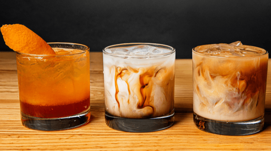 Three cocktails in short glasses on a wooden surface; the left drink is orange with a citrus peel, the middle and right drinks are creamy with visible swirling.