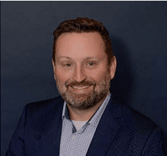 A man with a beard and short hair, wearing a blue blazer and a light patterned shirt, poses and smiles in front of a dark background.