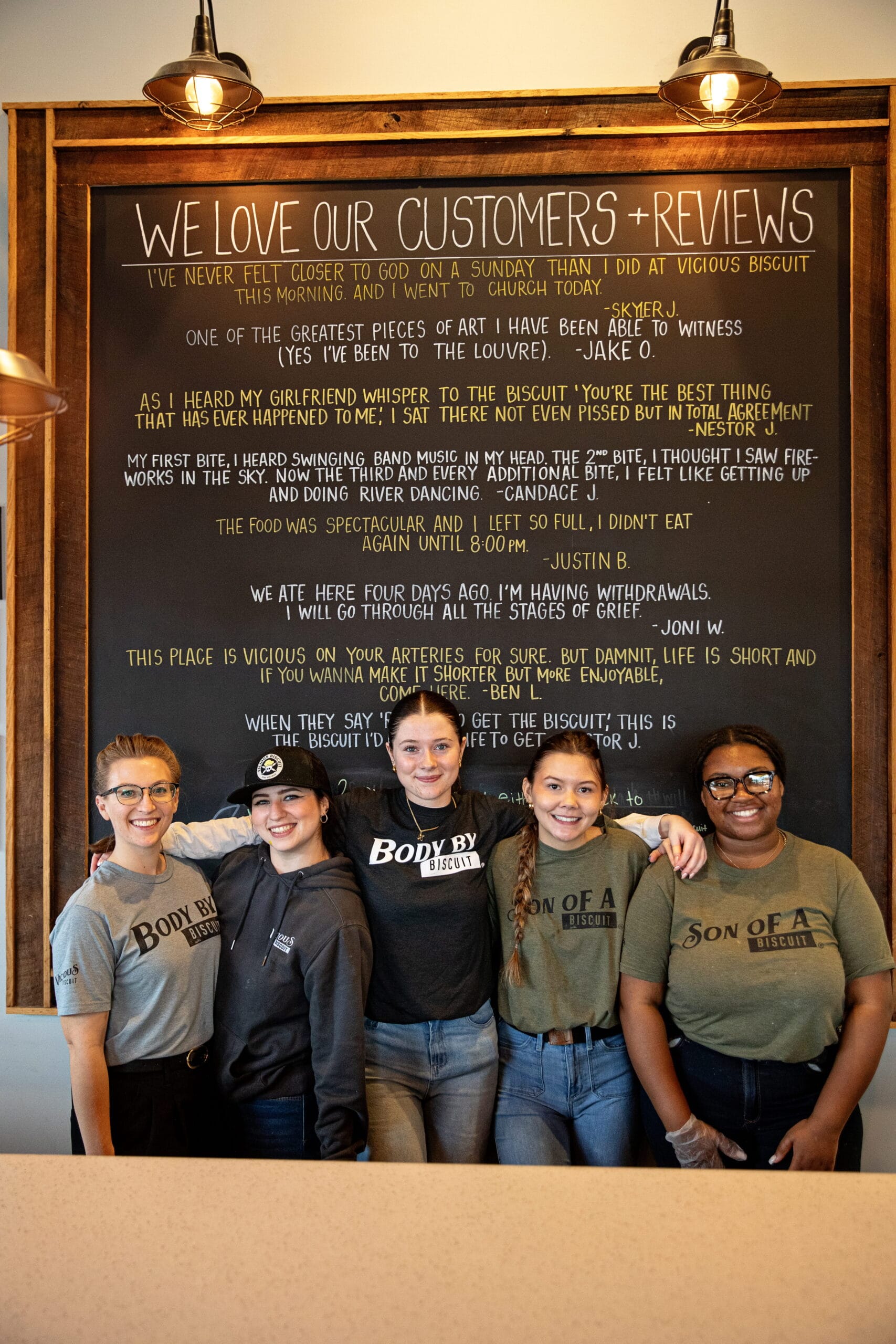 Five restaurant staff members stand smiling in front of a large chalkboard wall displaying customer reviews at a restaurant.