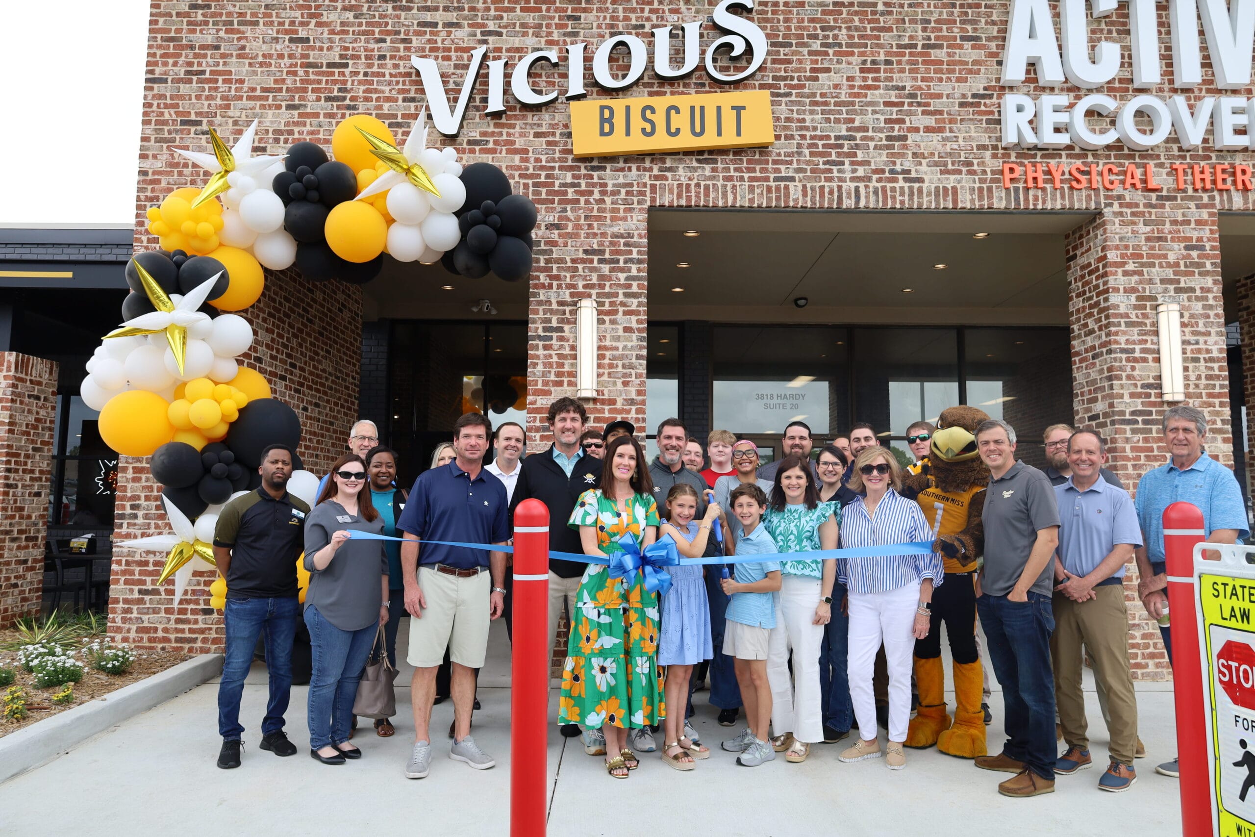 A group of people stand in front of Vicious Biscuit for a ribbon-cutting ceremony, with balloons decorating the entrance.