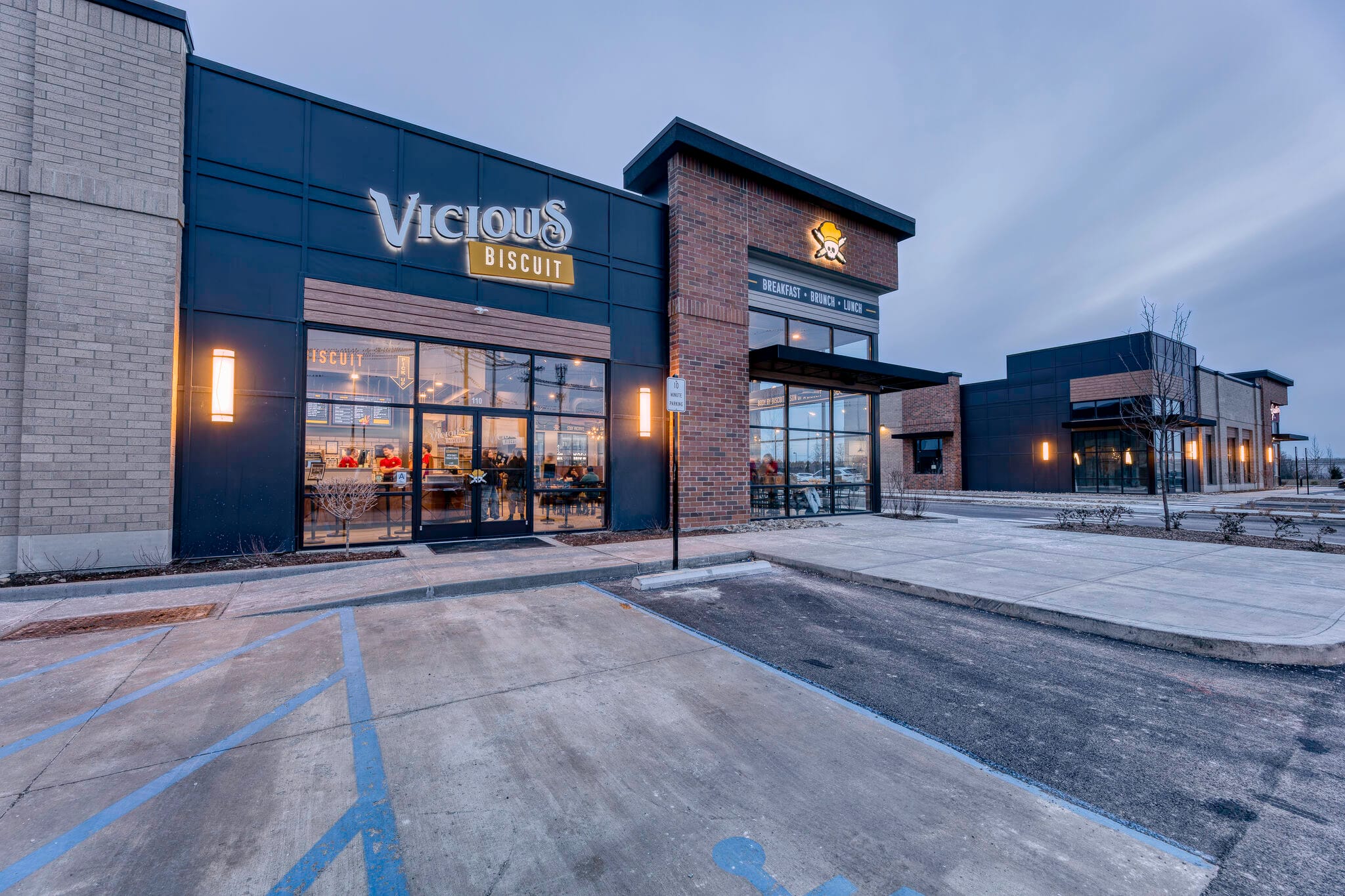 Exterior view of Vicious Biscuit restaurant with large glass windows, visible interior seating, and an adjacent empty parking lot at dusk.