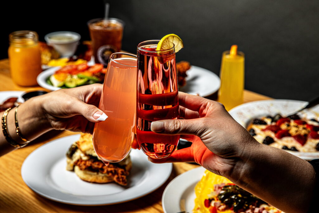 Two hands clink glasses with pink drinks over a table filled with assorted brunch foods, including eggs, fruit, and sandwiches.