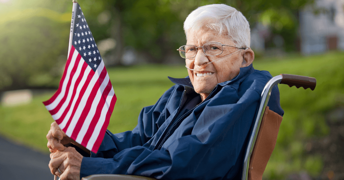 An older adult with glasses and a nasal cannula sits in a wheelchair outdoors, smiling and holding a small American flag.