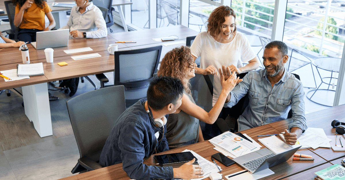 Four coworkers at a desk celebrate a successful project, high-fiving and smiling, with laptops, papers, and charts spread out in a modern office setting.
