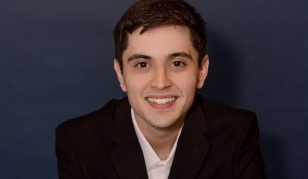 A young man with short dark hair, wearing a black blazer and white shirt, smiles at the camera against a plain dark blue background.