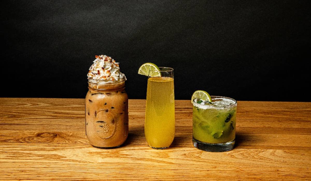 Three drinks on a wooden table: iced coffee with whipped cream in a jar, a yellow cocktail in a tall glass, and a green cocktail with lime in a short glass.